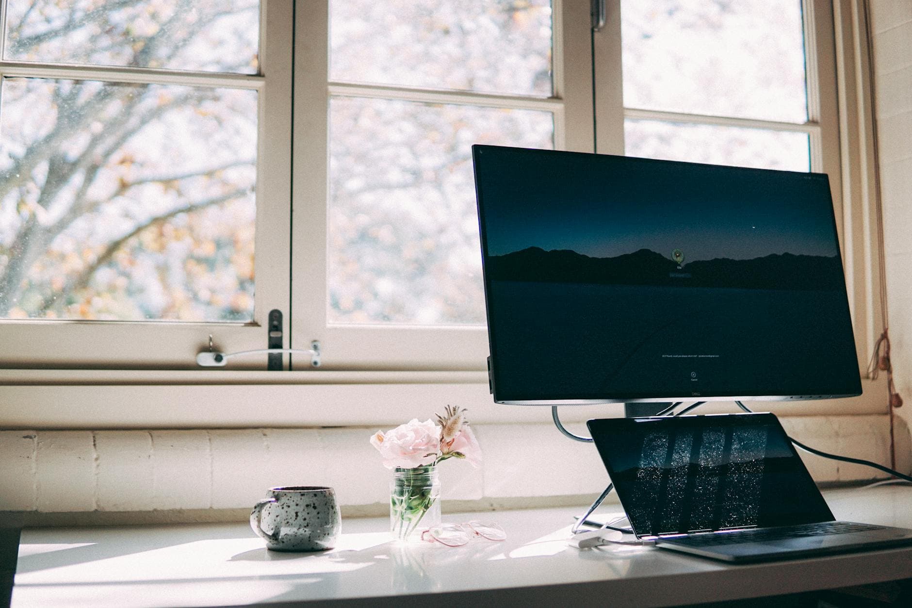 A calm home workspace near a bright window, with a laptop, a warm mug, and fresh flowers on the desk.