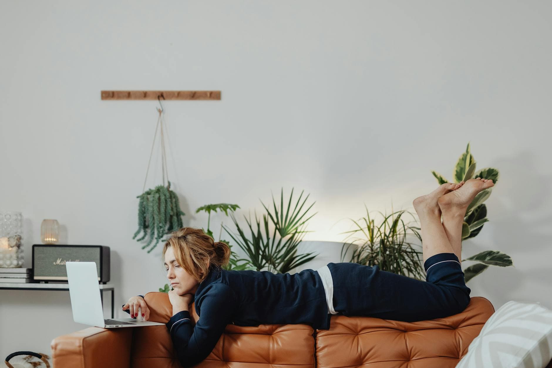 A woman lies on a couch working on her laptop at home, surrounded by indoor plants in a warm, calm living space.